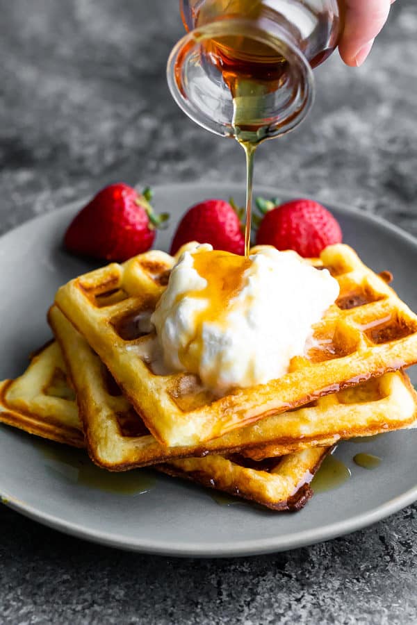 stack of three buttermilk waffles on a gray plate with syrup being drizzled over them and fresh strawberries
