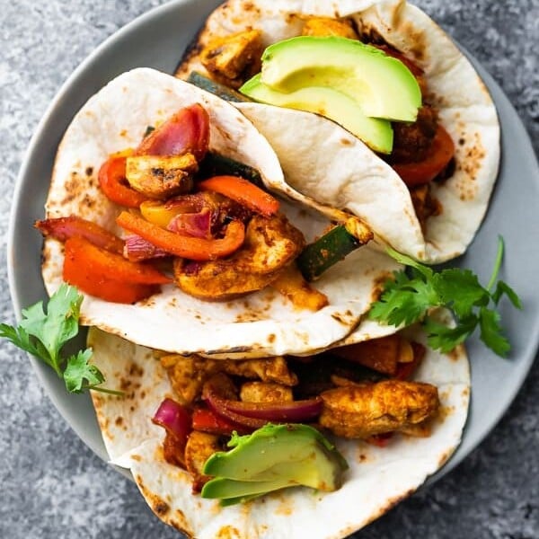 overhead shot of three sheet pan chicken fajitas on gray plate with fresh avocado and cilantro