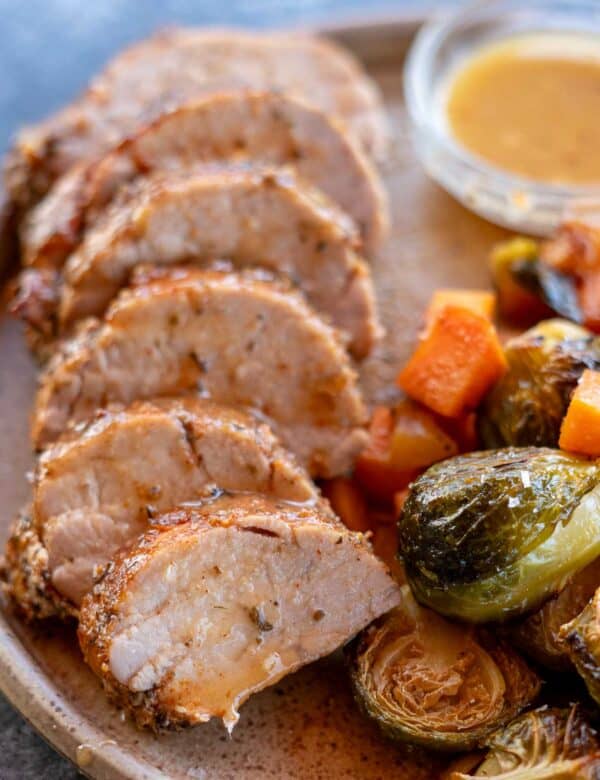 a closeup shot of a roasted pork tenderloin with juice on the plate and vegetables on the side