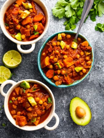 overhead shot of spicy slow cooker chickpea chili with white and blue bowls with fresh limes and avocado on gray background