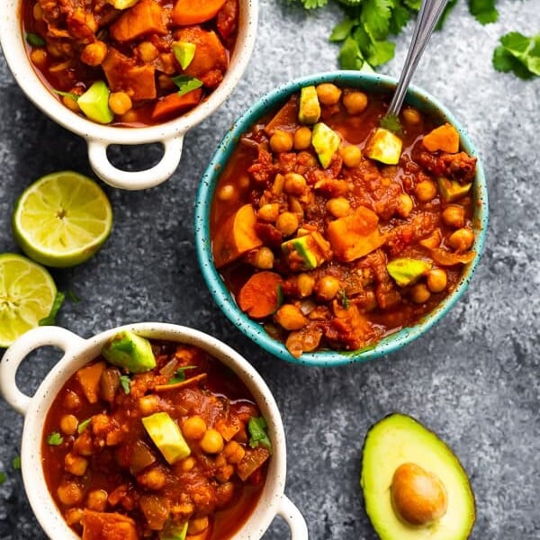 overhead shot of spicy slow cooker chickpea chili with white and blue bowls with fresh limes and avocado on gray background