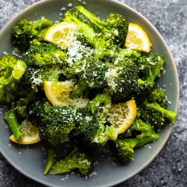 Overhead shot of a pile of broccoli with fresh lemon wedges and parmesan on a gray plate
