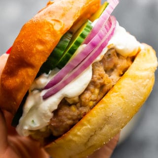 A hand holding a Greek Turkey burger in a bun with sliced red onion and cucumbers