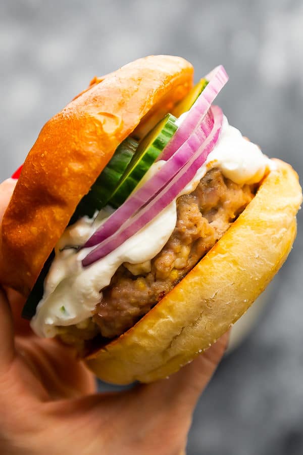 A hand holding a Greek Turkey burger in a bun with sliced red onion and cucumbers