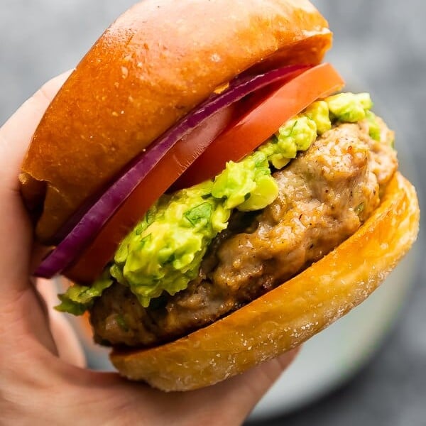 Hand holding a southwest turkey burger with guacamole, red onion slices, and tomato slices on a bun
