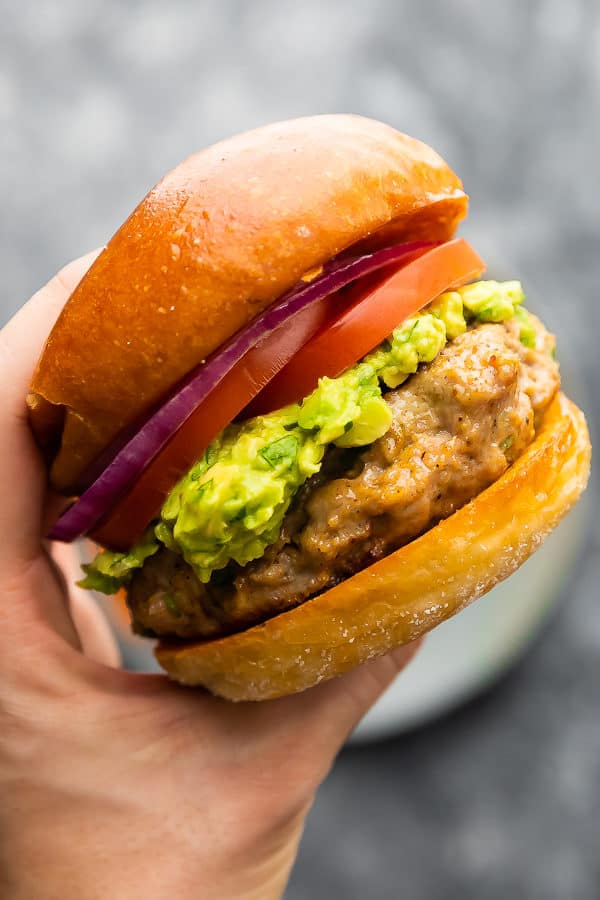 Hand holding a southwest turkey burger with guacamole, red onion slices, and tomato slices on a bun