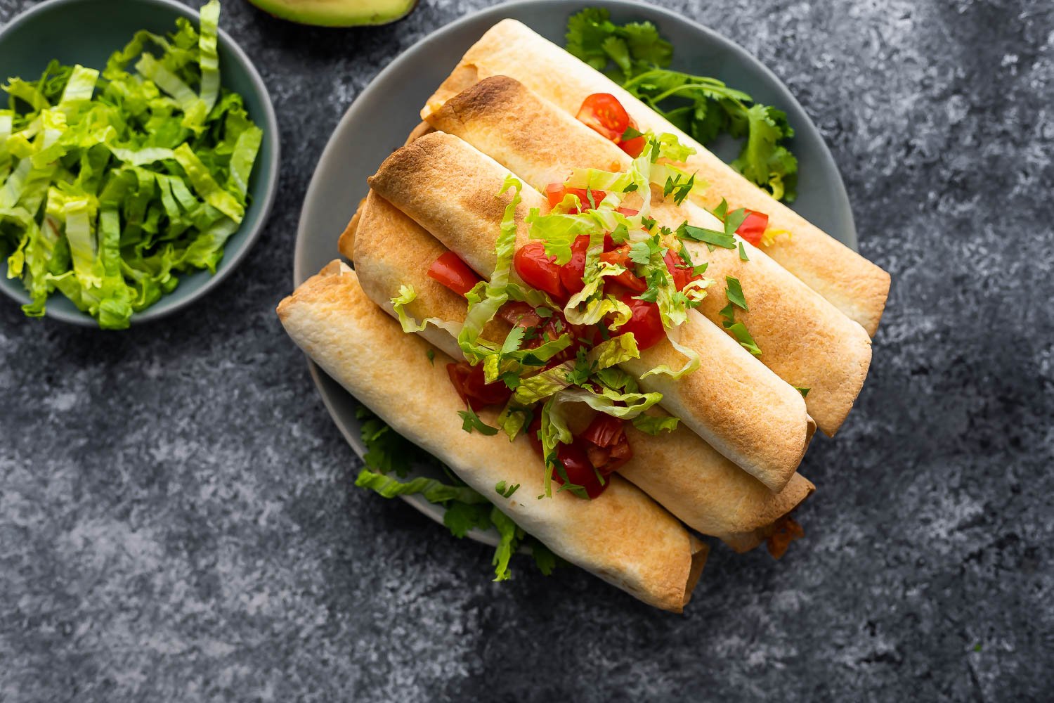 cooked taquitos stacked on a plate, topped with lettuce and tomatoes