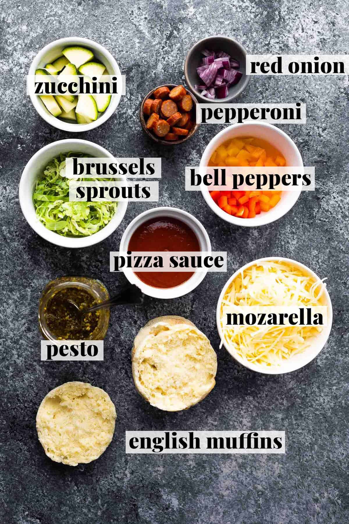ingredients for pizza in small ramekins on dark surface; including mozarella, bell peppers, pepperoni, red onion, zucchini, brussels sprouts, pesto, and english muffins