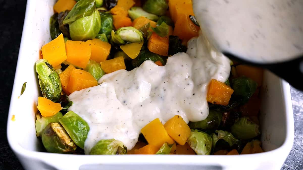 pouring cheese sauce over roasted brussels sprouts and butternut squash