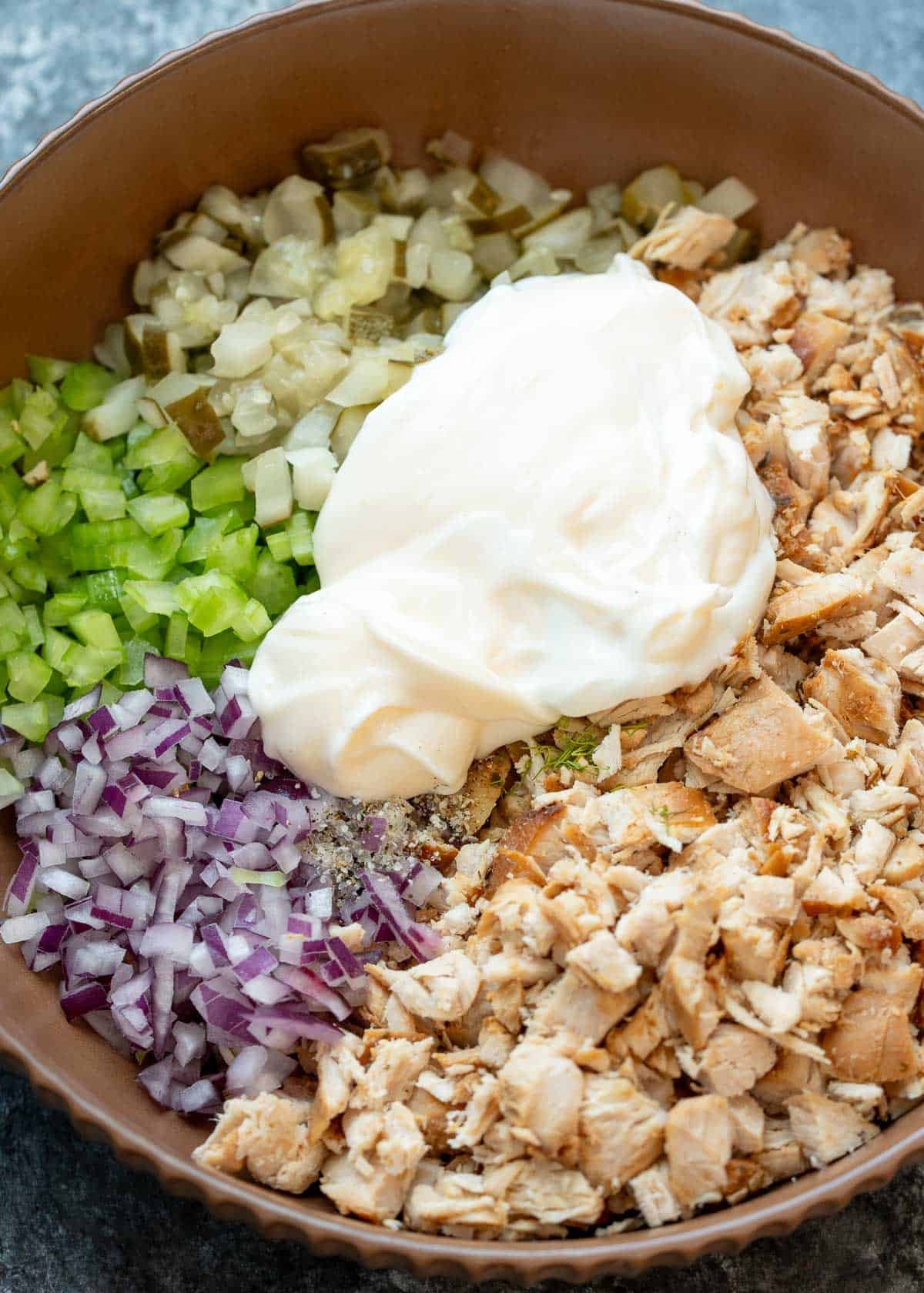mayonnaise, pickle juice, salt, and pepper added to chicken, celery, red onion, and pickles in a bowl for tangy chicken salad