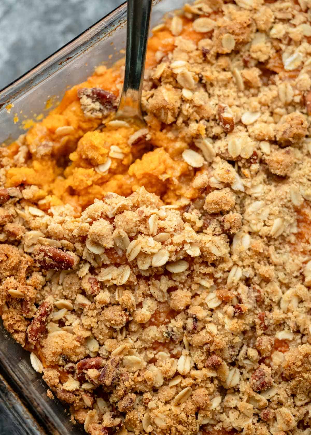 a closeup shot of a spoon scooping into a sweet potato casserole with brown sugar pecan topping