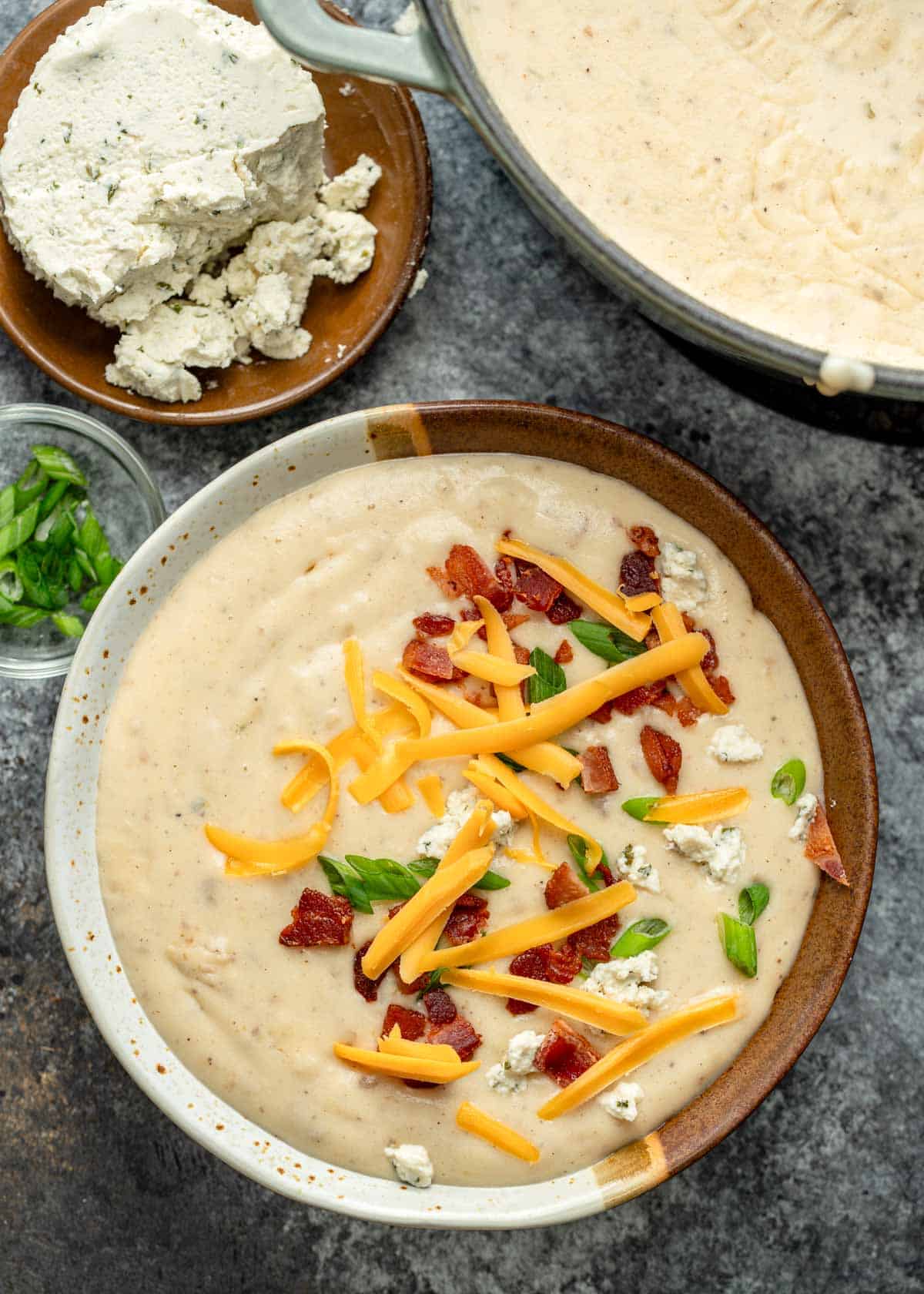 a bowl of boursin potato soup garnished with cheese, bacon, and green onions beside a bowl of boursin cheese and a pot of potato soup