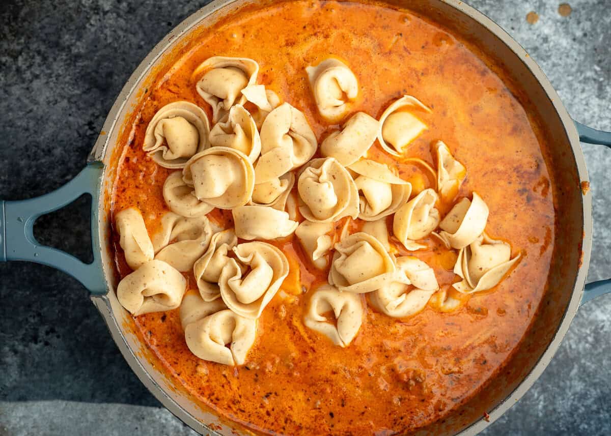 Tortellini being added to a creamy tomato meat sauce