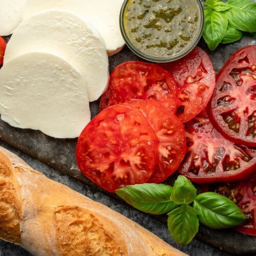 overhead image of sliced tomatoes, mozzarella, and a baguette on a stone cutting board