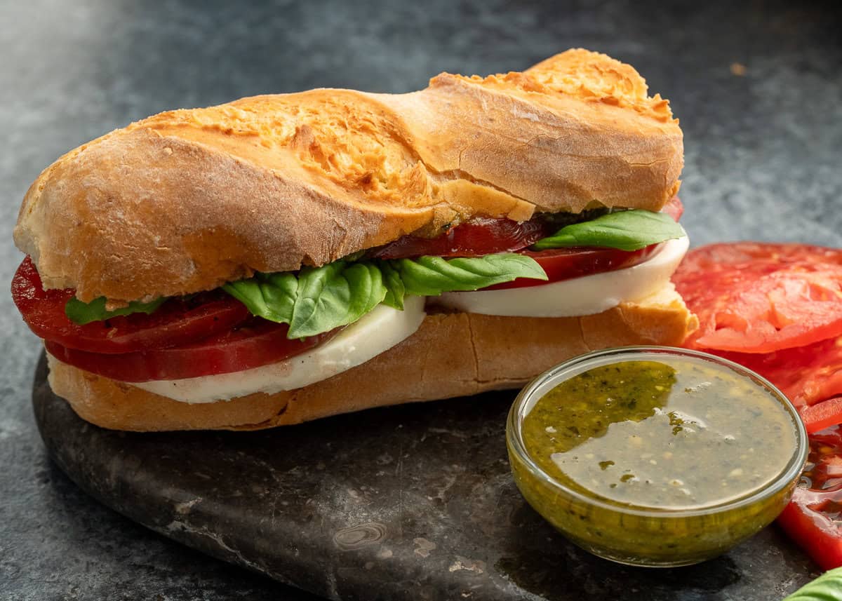 a caprese sandwich sitting with a jar of pesto on a stone cutting board