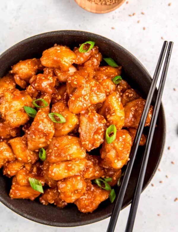 black bowl filled with sticky sesame chicken garnished with sesame seeds and green onions